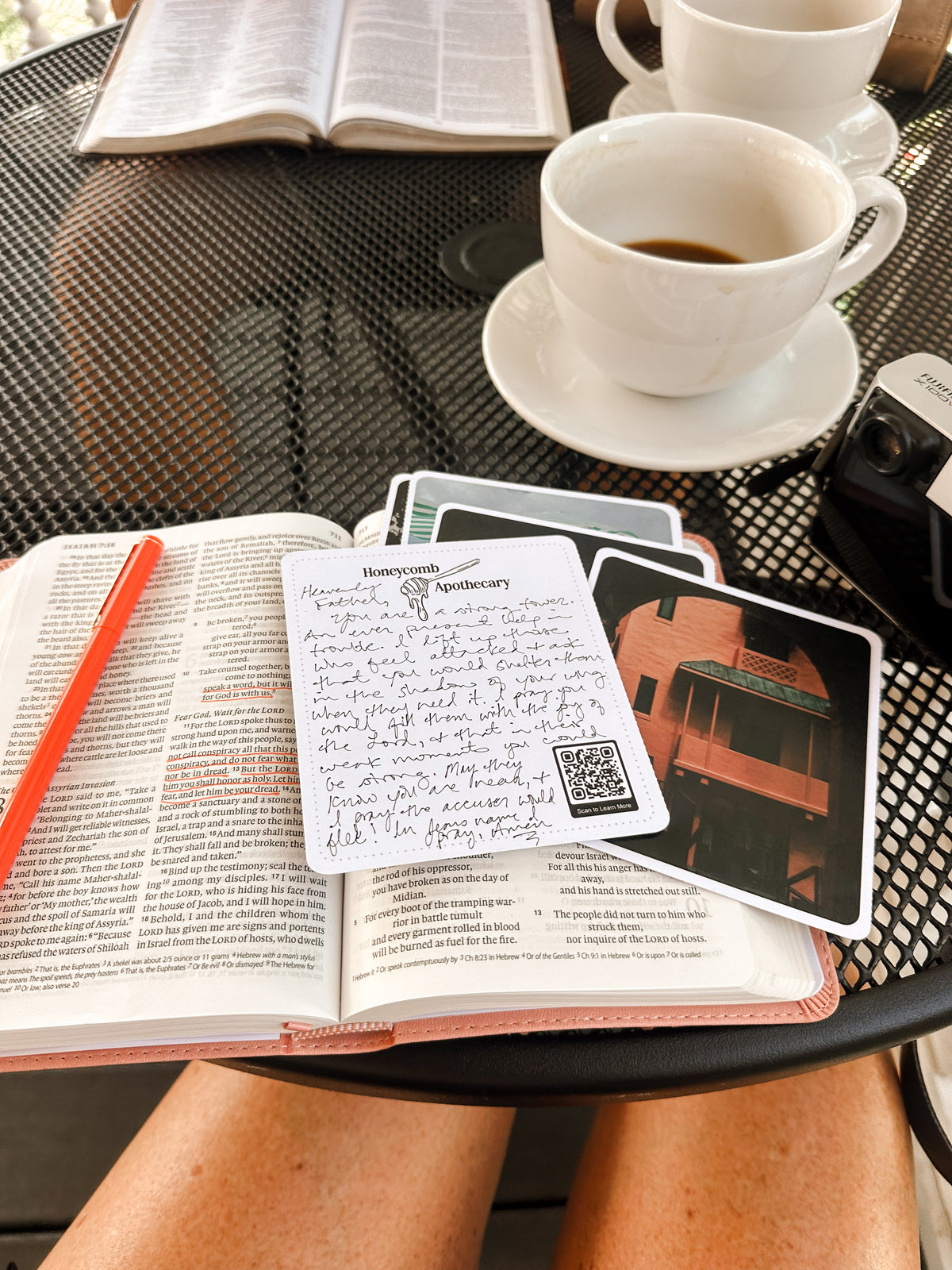 A person sitting at a black mesh outdoor table with an open Bible, a pink pen, handwritten cards, and coffee cups on the table.